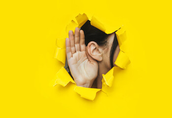 Wall Mural - Close-up of a woman's ear and hand through a torn hole in the paper. Bright yellow background, copy space. The concept of eavesdropping, espionage, gossip and tabloids.
