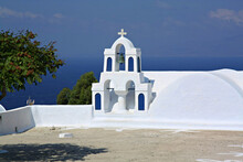 White Clock Tower With A Cross By The Sea On Santorini, Island Greece With Blue Sky On The Horizon
