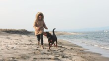 A Girl In A Jacket Runs And Fools Around On The Beach By The Sea With A Rottweiler Dog