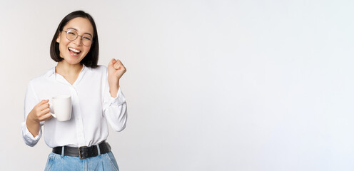 Happy dancing woman drinking coffee or tea from mug. Korean girl with cup, standing over white background