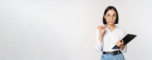 Wall Mural - Asian girl in glasses thinks, holds pen and clipboard, writing down, making notes, standing over white background