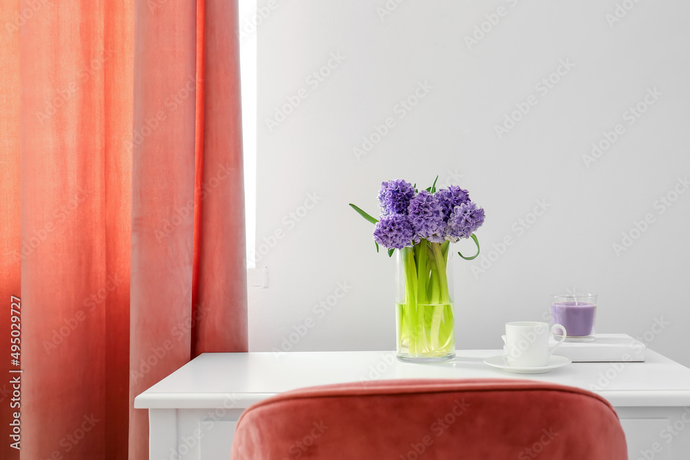 Vase with hyacinth flowers, cup and candle on table near light wall