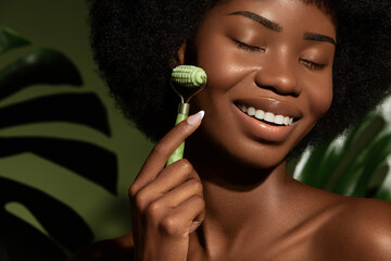 Wall Mural - gua sha face care. african american young woman massaging her face skin with jade roller against gre