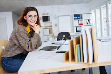 Wall Mural - young business woman sits at a desk with laptop and laughs into the camera