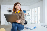 Fototapeta Panele - young happy business woman sitting on a desk with laptop in her hands