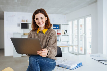Wall Mural - young smiling business woman sitting on a desk with laptop in her hands