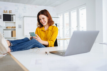 Wall Mural - young modern business woman sitting relaxed at her desk and looking at her cell phone in the office