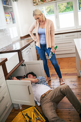 Poster - Honey, I think we should just call a professional. Shot of a man fixing pipes under his kitchen sink while his wife watches.