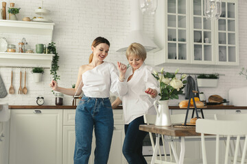 Elderly woman dancing to energetic music with grown up daughter. Happy smiling two generations female family having fun together in kitchen