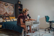 Portrait Photo Of A Young Teenage Boy In Headphones Recording Voice Or Chatting With Friends Using A Microphone With Pop Filter And Laptop Sitting On A Bed In His Home Room..