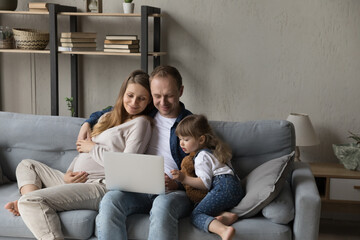 Happy young bonding father and pregnant mother using computer applications with adorable little child daughter, watching cartoons or comedian movie online, enjoying playing games or shopping.