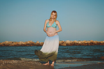 Wall Mural - Beautiful young elegant pregnant blond woman in a blue hat walks on the beach, seashore at sunset. Azov sea. Motherhood. Maternity.