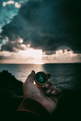 Wall Mural - Pov of man hand holding navigational travel compass to find direction and next destination place. Concept of travel people and exploring. Beach and ocean with sdramatic ky light in background