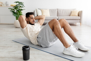 Wall Mural - Full length of sporty young Arab man strengthening core muscles, doing abs exercises at home