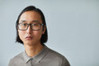© Seventyfour - Minimal close up portrait of young Asian businessman wearing glasses and looking at camera while standing against light blue background in studio, copy space