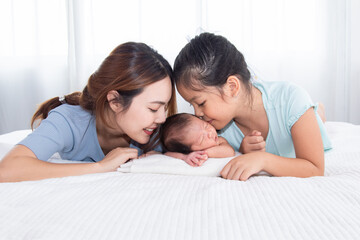 Beautiful Asian mother and daughter spend time with newborn in bed, two woman kiss adorable infant with love and care. Toddler baby sleeping in bedroom with family.