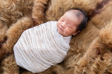 Asian little newborn baby deeply sleeping wrapped in thin white clothwith happy and safe on brown wool blanket. adorable infant lying on his back relaxing under a white color cloth.