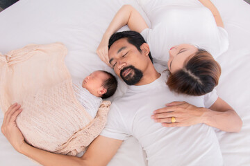 Wall Mural - Asian young mother father and newborn sleep on bed together. Parent take a nap with baby because tired nurturing baby. Little infant  deeply sleeping wrapped in thin white cloth with happy and safe.