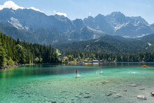 The German Mountain Lake Eibsee With Clear Water And People On Their Stand Up Paddle Boards With The Mountain Zugspitze In The Background.