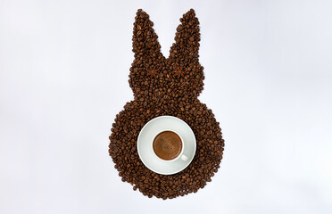 Creative top view flat lay of Easter bunny shape made of coffee roasted beans on white background. Rabbit is decorated with anise star seeds as eyes and cup of espresso as tail. 