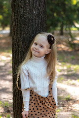 Wall Mural - Portrait of a blonde girl in a beige turtleneck and skirt. The girl is standing on a tree. Childhood. Tenderness.