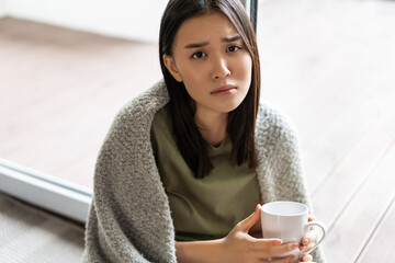 Wall Mural - Sad and gloomy asian woman sitting at home near window, holding cup of coffee and frowning disappointed, feeling unhappy or miserable