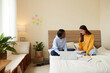 © DragonImages - Smiling young woman explaining difficult topic to friend when they are sitting on bed in dormitory