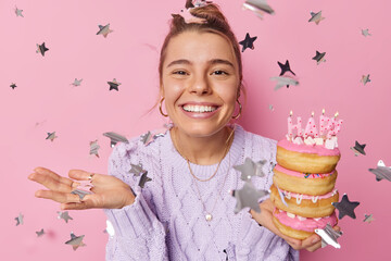 Wall Mural - Happy young European woman with combed hair toothy smile holds pile of sweet doughnuts with burning candles poses against pink background flying stars around celebrates birthday waits for guests