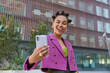 © wayhome.studio  - Fashionable young woman with bun hairstyle winks eye poses for selfie at smartphone camera wears fashionable pink jacket poses outdoors in urban setting has cheerful expression. Street lifestyle
