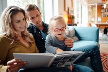 Father And Mother Looking At Cute Daughter Reading Book In Living Room At Home