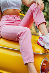 Stylish woman with pretty legs in pink fashionable jeans with colored sneakers sitting on a yellow vintage car. Women's summer bright fashion style.