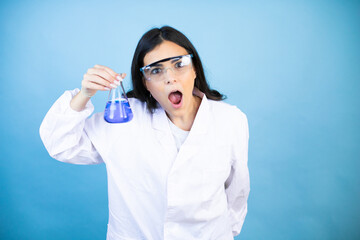 Wall Mural - Young brunette woman wearing scientist uniform holding test tube over isolated blue background afraid and shocked with surprise and amazed expression, fear and excited face.