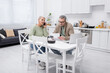 © LIGHTFIELD STUDIOS - Elderly woman talking on smartphone while husband holding paper near coffee and laptop in kitchen.