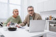© LIGHTFIELD STUDIOS - Pensioners looking at laptop and holding papers near coffee in kitchen.