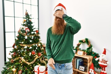 Poster - Redhead man with long beard wearing christmas hat by christmas tree covering eyes with hand, looking serious and sad. sightless, hiding and rejection concept