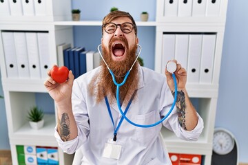 Canvas Print - Redhead man with long beard wearing doctor uniform holding heart and stethoscope angry and mad screaming frustrated and furious, shouting with anger looking up.