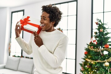 Sticker - Young african american man holding gift standing by christmas tree at home.