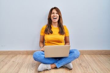 Sticker - Hispanic woman using laptop sitting on the floor at home angry and mad screaming frustrated and furious, shouting with anger. rage and aggressive concept.