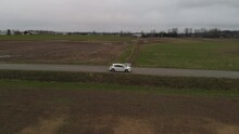 White Car 2019 Toyota Corolla Hatchback Driving Along Country Farm Road Surrounded By Grass Brown Fields Mountains In Farmland Abbotsford BC Wide Aerial Side Profile Tracking As Car Accelerates