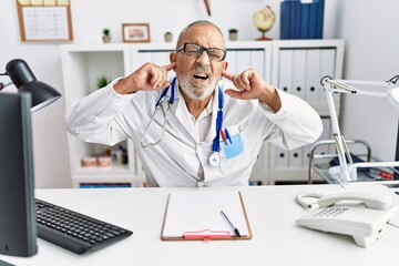 Poster - Mature doctor man at the clinic covering ears with fingers with annoyed expression for the noise of loud music. deaf concept.