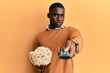 Young african american man holding television remote control eating popcorn relaxed with serious expression on face. simple and natural looking at the camera.