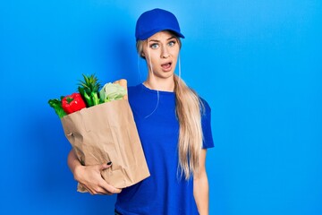 Wall Mural - Young caucasian woman wearing courier uniform with groceries from supermarket in shock face, looking skeptical and sarcastic, surprised with open mouth