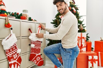 Canvas Print - Young hispanic man smiling happy wearing christmas hat holding gift at home.