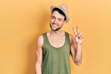 Canvas Print - Young hispanic man wearing summer hat smiling with happy face winking at the camera doing victory sign. number two.