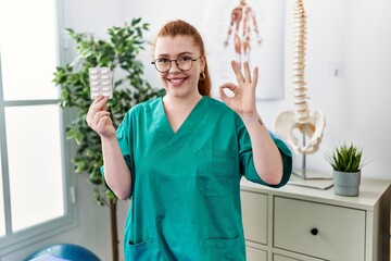 Canvas Print - Young redhead physiotherapist woman holding prescription pills doing ok sign with fingers, smiling friendly gesturing excellent symbol