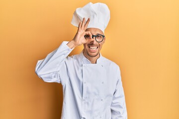 Bald man with beard wearing professional cook uniform doing ok gesture with hand smiling, eye looking through fingers with happy face.