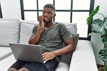 Sticker - Young african american man using laptop at home sitting on the sofa looking stressed and nervous with hands on mouth biting nails. anxiety problem.