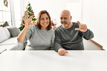 Wall Mural - Middle age hispanic couple sitting on the table by christmas tree smiling and confident gesturing with hand doing small size sign with fingers looking and the camera. measure concept.