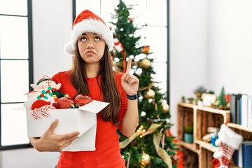 Sticker - Young brunette woman standing by christmas tree holding decoration pointing up looking sad and upset, indicating direction with fingers, unhappy and depressed.