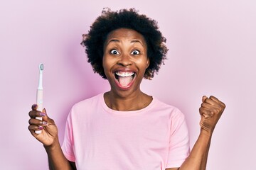 Sticker - Young african american woman holding electric toothbrush screaming proud, celebrating victory and success very excited with raised arms